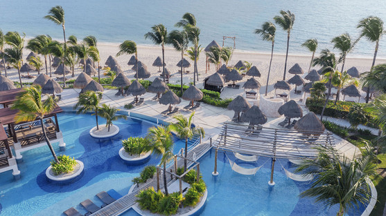 An aerial view of the tropical pool complex at Excellence Playa Mujeres in Mexico, featuring winding turquoise pools with built-in lounge areas and palm-tree islands. The resort's white-sand beach is lined with numerous thatched-roof palapas and swaying palms, leading to the calm waters of the Caribbean Sea under a clear sky.