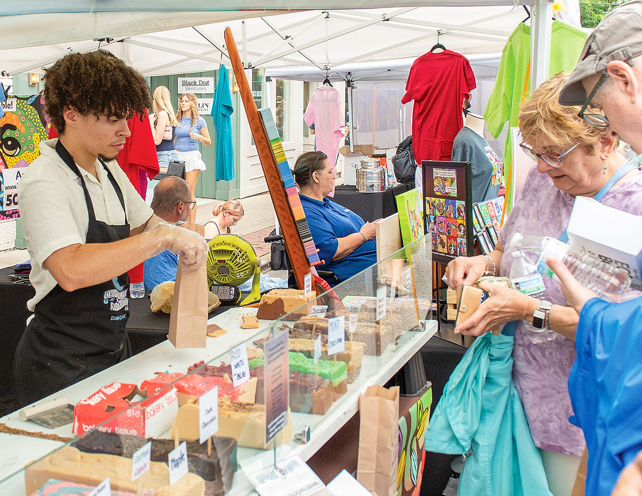 A vendor works behind a table displaying various sweet treats and fudge while customers shop at the outdoor market.