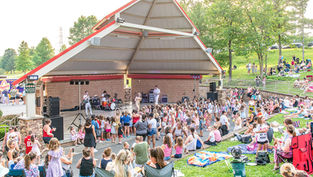 A wide-angle shot shows an outdoor amphitheater with a covered stage where a live band, including the Taylor Swift performer, is playing. A large crowd of children is gathered directly in front of the stage, while families sit on blankets and in lawn chairs on the grassy hill behind them. The sun is setting, casting a warm glow on the trees and the crowd.