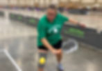 An action shot of a female pickleball player indoors, wearing a green shirt and black shorts, demonstrating a backhand serve technique.