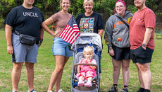 A group of five adults stand together on a grassy field, smiling at the camera, with a toddler in a pink outfit sitting in a stroller in the center, holding a small American flag.