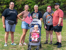 A group of five adults stand together on a grassy field, smiling at the camera, with a toddler in a pink outfit sitting in a stroller in the center, holding a small American flag.
