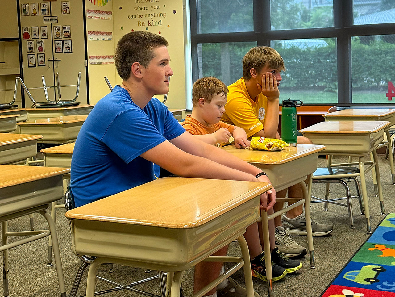 Three boys sit at desks in a classroom, listening to someone off-camera.