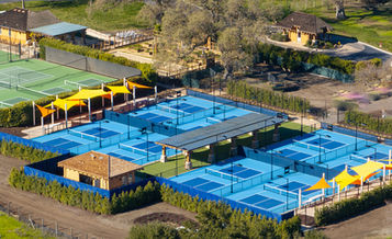An aerial view of a complex of multiple blue pickleball courts at CordeValle. The courts have orange and yellow sun shades and are surrounded by lush greenery, a fence, and a building with a tile roof.