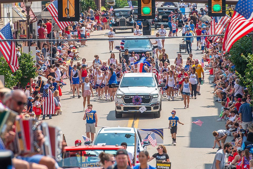 An aerial view of a vibrant Fourth of July parade with a crowd of people watching from the sidewalks and many floats and marchers in the street.