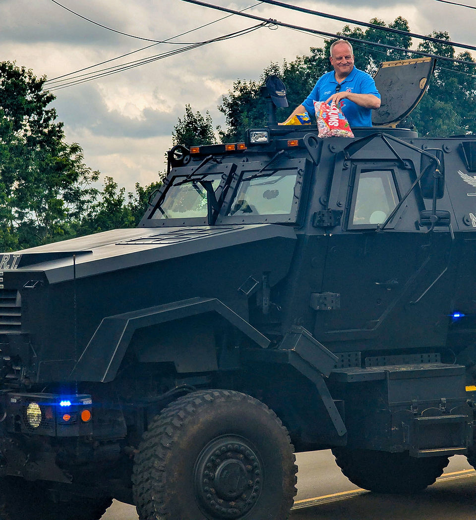 A low-angle, close-up photograph of a large, black, military-style armored tactical vehicle moving down a street. The vehicle has large, knobby tires, multiple armored windows, and several blue and yellow flashing emergency lights. A man with short grey hair, wearing a blue polo shirt, is standing up through an open roof hatch, smiling and looking towards the right. He is holding a small bag of chips and seems to be greeting people along the parade route. The background shows green trees and grey, cloudy skies, with some power lines visible. The front of the vehicle has an "R.T." marking.