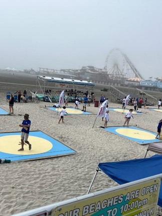 Misty aerial view of the National Marbles Tournament on a beach in Wildwood, New Jersey, showing several square blue and yellow marble rings set up on the sand, with the famous boardwalk and amusement rides visible in the background.