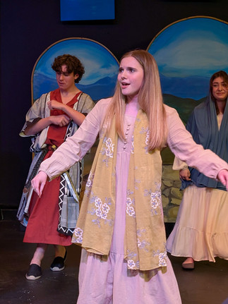 A medium shot of Sylvia Youshack performing on stage in the role of Mary. She is wearing a light purple, long-sleeved dress paired with a yellow floral-patterned scarf. She has a focused, emotional expression as she sings with her arms slightly outstretched. In the background, other cast members in biblical robes stand before the painted blue mountain backdrops.
