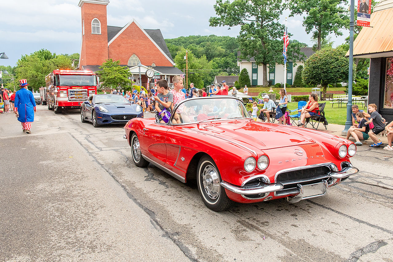 A vintage red convertible Chevrolet Corvette driving down a town street during a Fourth of July parade, carrying two young boys waving to spectators, followed by a sleek blue sports car, a bright red fire truck, and a person walking in an Uncle Sam costume.