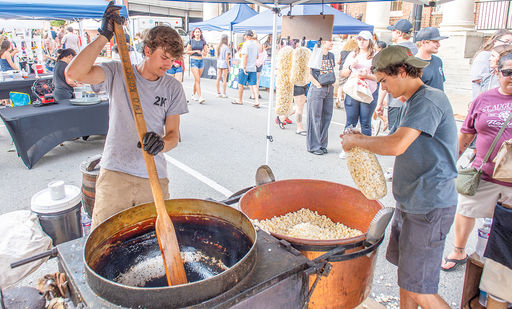 Two young men operate a kettle corn stand at an outdoor market. The man on the left, wearing a grey t-shirt and khaki pants, actively stirs a large, dark, hot kettle with a long wooden paddle. The man on the right, wearing a dark t-shirt, shorts, and a backwards baseball cap, holds a large clear plastic bag, filling it with freshly popped kettle corn from a second, copper-colored kettle. Both are under a white canopy with other market stalls and attendees visible in the background.