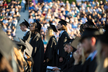 A sunlit, side-angle view of a row of high school graduates standing in unison during the outdoor ceremony. They are wearing black caps and gowns, looking attentively toward the stage. In the bright, out-of-focus background, large stadium bleachers are packed with a crowd of spectators.