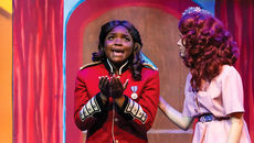 A young actress in a red military-style jacket with gold epaulettes stands center stage with her hands clasped in a pleading gesture, looking upward with an emotional expression. To her right, another actress in a pink dress and a floral headpiece places a supportive hand on her shoulder.