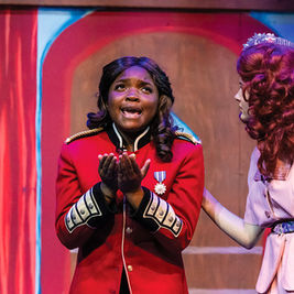 A young actress in a red military-style jacket with gold epaulettes stands center stage with her hands clasped in a pleading gesture, looking upward with an emotional expression. To her right, another actress in a pink dress and a floral headpiece places a supportive hand on her shoulder.