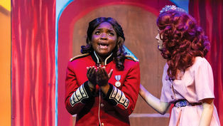 A young actress in a red military-style jacket with gold epaulettes stands center stage with her hands clasped in a pleading gesture, looking upward with an emotional expression. To her right, another actress in a pink dress and a floral headpiece places a supportive hand on her shoulder.