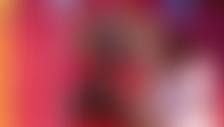 A young actress in a red military-style jacket with gold epaulettes stands center stage with her hands clasped in a pleading gesture, looking upward with an emotional expression. To her right, another actress in a pink dress and a floral headpiece places a supportive hand on her shoulder.