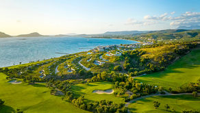 An elevated, wide-angle photograph of a luxurious, modern coastal resort complex, presumably Navarino Dunes. The resort features low-profile, sand-colored buildings with green, landscaped roofs, nestled into a hillside that drops down to a turquoise bay. In the foreground and sweeping around the resort is a sprawling, vibrant green golf course, complete with bunkers and olive trees. In the distance across the water, other landmasses are visible under a clear sky with scattered clouds, bathed in the golden light of a setting or rising sun.