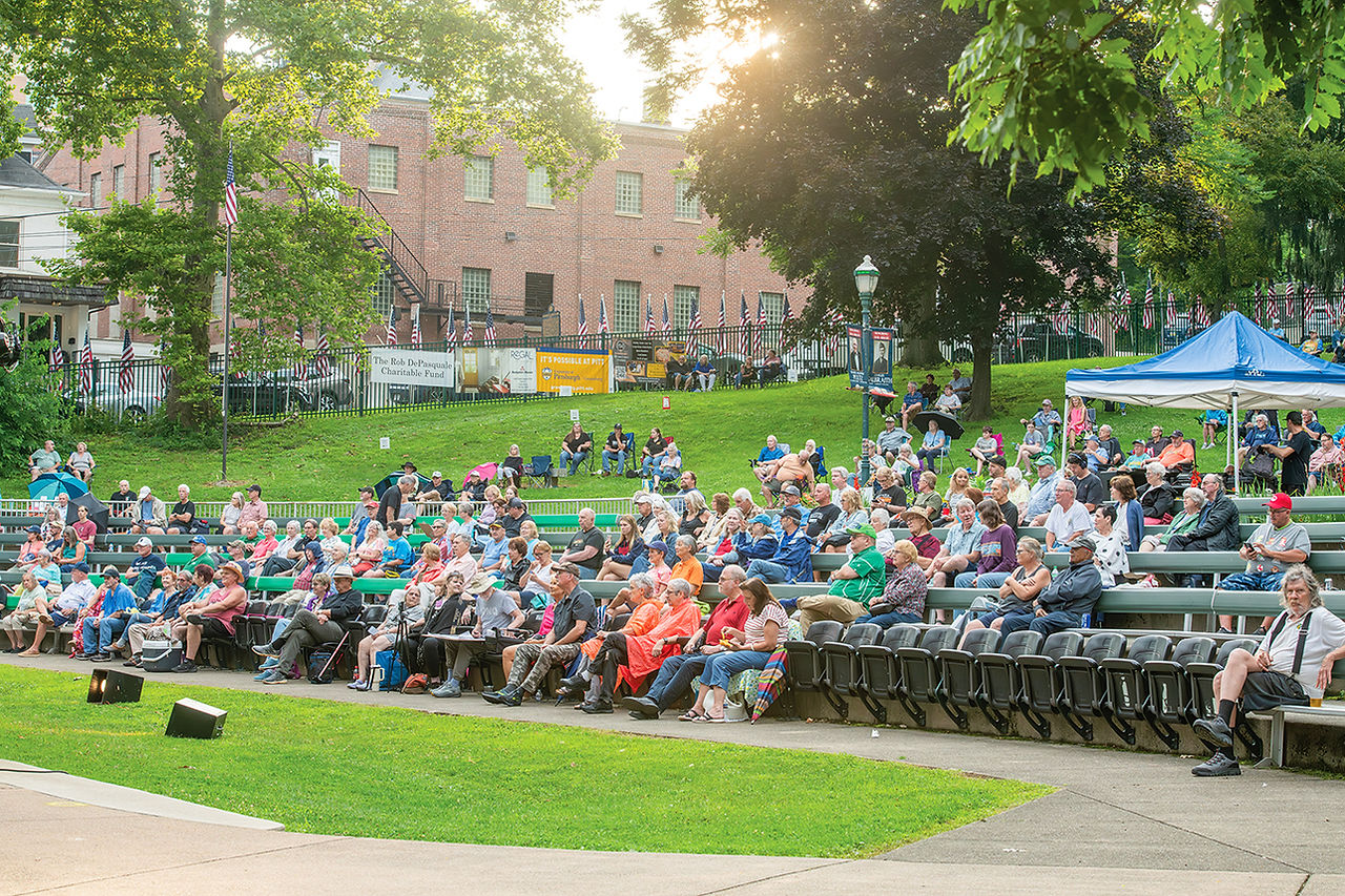 A wide-angle view of an outdoor amphitheater during a sunny late afternoon concert. Rows of green metal bleachers are filled with an audience of adults and seniors seated and enjoying the music. A blue pop-up canopy tent stands on the right side of the seating area. In the background, a terraced grassy hill rises behind a fence adorned with numerous small American flags, leading to a brick building and mature trees bathed in warm sunlight.