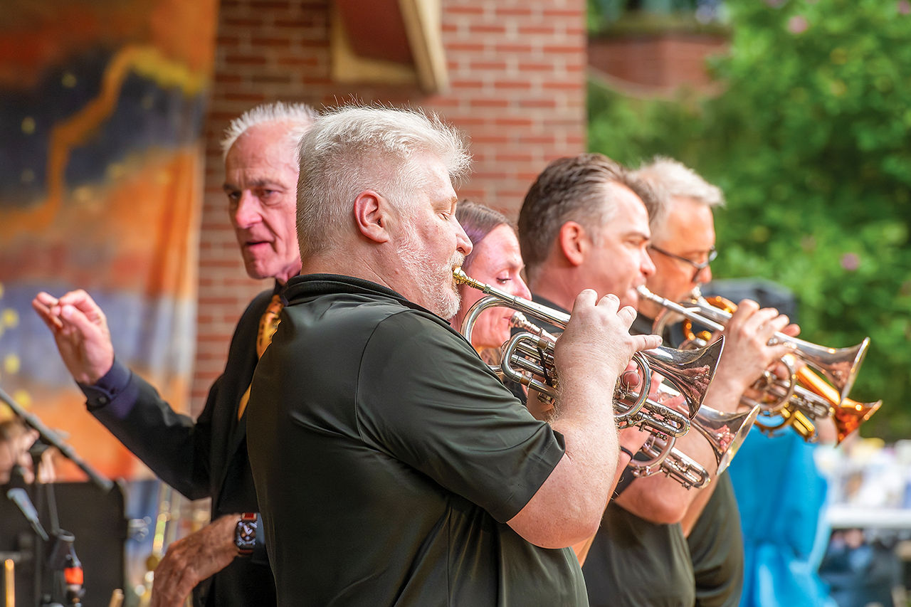 A side-profile shot of four cornet and trumpet players performing on stage. In the foreground, a musician with silver-white hair and a trimmed beard plays a silver cornet. Behind and beside him, other band members in dark polo shirts play their instruments with focused expressions. To the far left, a conductor or featured performer in a dark suit jacket and tie raises a hand to cue the music. The background features a brick wall and a portion of a warm, abstract stage backdrop.
