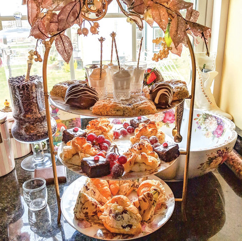 A close-up photograph of a three-tiered golden-metal tea stand, laden with a variety of afternoon tea treats. The top tier holds individual cups of mousse, scones, and chocolate-dipped fruit. The middle tier features delicate pastries filled with fresh berries, and the bottom tier is piled with assorted finger sandwiches. The background shows a table next to a sunny window with frilly curtains.