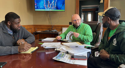 Mayor Tom Maglicco (center), wearing a bright green quarter-zip sweater and glasses, leads a meeting around a polished wooden conference table. He is flipping through a colorful brochure while speaking to two men. The man on the left, in a grey zip-up jacket, takes notes on a yellow legal pad. The man on the right wears a green "Local 6" hoodie and a camouflage hat. The table is covered in various documents, maps, and architectural renderings, with a large television screen displaying a blue abstract image in the background.