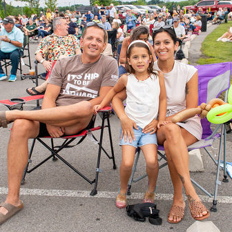 A family of three sitting together in folding chairs at an outdoor park event, with the young daughter holding a balloon animal.