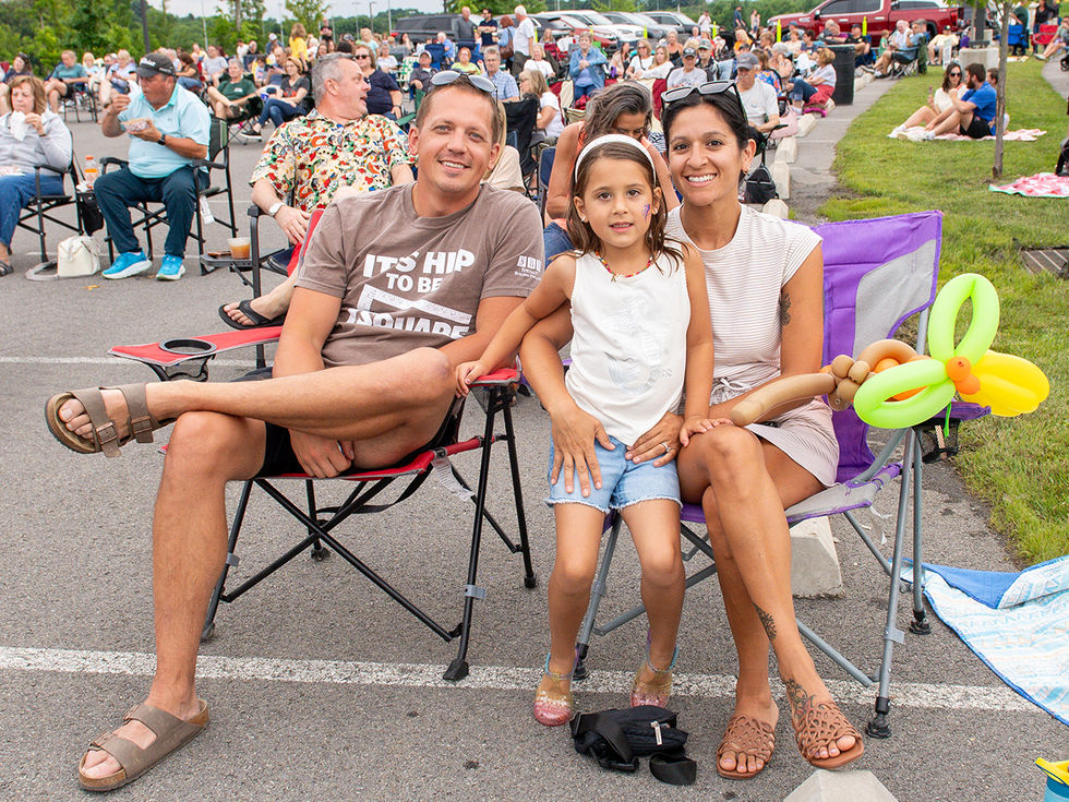 A family of three sitting together in folding chairs at an outdoor park event, with the young daughter holding a balloon animal.