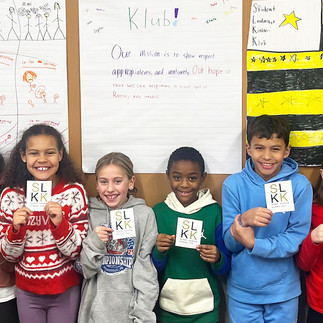 A group of eight elementary students from Ramsey Elementary stand together, each holding a square "SLKK" (Student Leadership Kindness Klub) card. They are positioned in front of hand-drawn posters, including one outlining their mission to show respect and maturity and another featuring a hand-drawn American flag with yellow stars and stripes. The students are smiling, showcasing their role as school role models.
