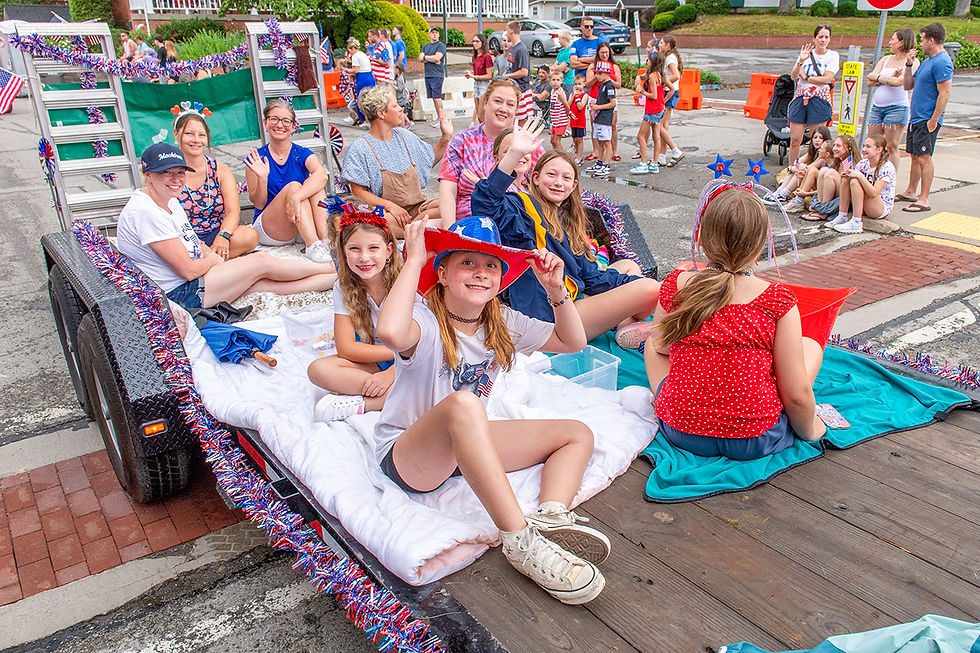 A close-up photograph taken from the back of a moving flatbed trailer during a parade. Several women and children are seated on white bedding, waving towards the crowd on the sidewalk. The trailer's metal rails are decorated with red, white, and blue tinsel. A girl in a red hat is prominent in the foreground. In the background, spectators, including a woman in a white shirt and blue shorts, watch from the sidewalk. A yellow truck is visible further down the street.