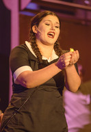 This shot focuses on a singing woman who is center stage. She is singing and her mouth is wide open, with a microphone clearly visible on her right cheek. She has her hair parted in the middle and braided in two tight braids, and she is wearing a black-and-white maid uniform. Her dark reddish-brown lipstick complements the rich purple and pink lighting. She is holding something small and yellow in both hands. The backdrop, a slightly out-of-focus mix of pink and light purple, contrasts the woman.