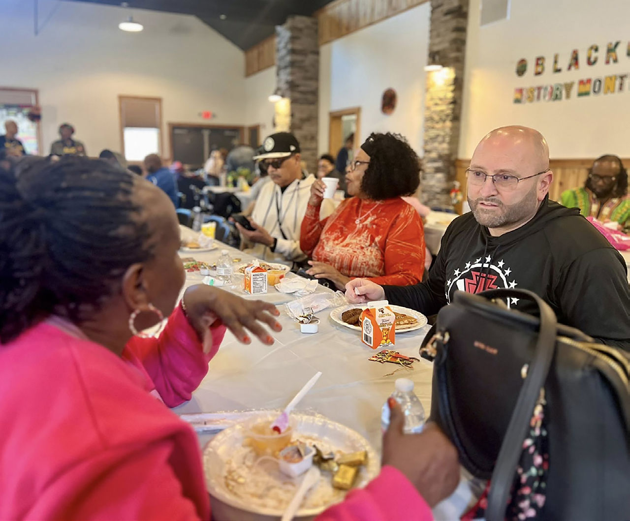 Mayor Tom Maglicco (right), wearing a black hooded sweatshirt with a circular red and white logo, is seated at a long banquet table covered in a white cloth. He is engaged in conversation with several community members during a Black History Month breakfast event. In the foreground, a woman in a bright pink top is partially visible, and in the background, other attendees are eating and talking. A "Black History Month" sign is visible on the wall behind the table.