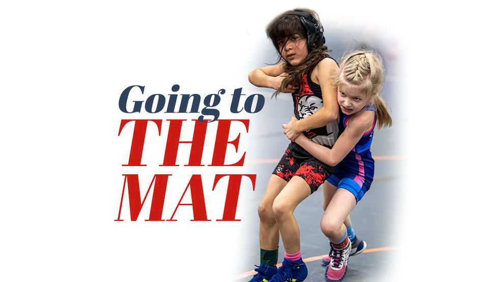 An action shot of Maya Bhandari in a red and black wrestling singlet and headgear, engaged in a competitive match. She is being held from behind by an opponent in a blue singlet as they both grapple for position on a grey wrestling mat. To the left, the stylized article title "Going to the MAT" is overlaid in bold blue and red text.