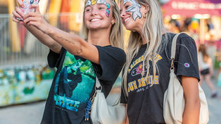 Two smiling young women taking a selfie; both have fun face painting designs, one with a shark and one with a rainbow unicorn horn.