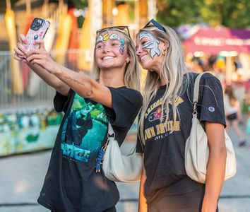 Two smiling young women taking a selfie; both have fun face painting designs, one with a shark and one with a rainbow unicorn horn.