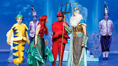 On a stage, a performer dressed as Ariel speaks with a performer dressed as King Triton, who holds a trident, while other cast members in colorful aquatic-themed costumes stand nearby against a blue, underwater-themed backdrop.