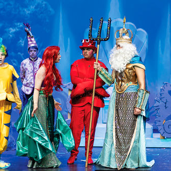 On a stage, a performer dressed as Ariel speaks with a performer dressed as King Triton, who holds a trident, while other cast members in colorful aquatic-themed costumes stand nearby against a blue, underwater-themed backdrop.