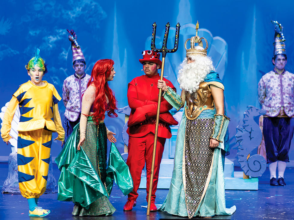 On a stage, a performer dressed as Ariel speaks with a performer dressed as King Triton, who holds a trident, while other cast members in colorful aquatic-themed costumes stand nearby against a blue, underwater-themed backdrop.