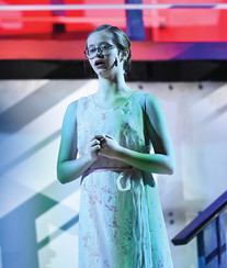 A student performs a solo moment in Greensburg Salem High School’s spring musical Anything Goes, standing center stage in a light floral dress with her hands clasped near her chest. Cool blue-green lighting falls across her as part of the ship-themed set and staircase appear behind her, creating a quiet, reflective stage moment.
