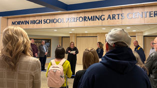 A view from behind a small crowd of attendees gathered in the lobby of the new Norwin High School Performing Arts Center. A woman in a black dress stands at the center, speaking to the guests and holding a piece of paper. The entrance to the theater is marked by large blue lettering on wood paneling that reads "NORWIN HIGH SCHOOL PERFORMING ARTS CENTER." Several district officials stand to the side, listening to the presentation.