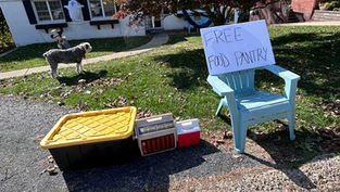 A photograph showing the humble beginnings of a community food pantry on a residential driveway. A large black plastic storage bin with a bright yellow lid sits next to a small vintage cooler and a red Playmate cooler. A light blue plastic Adirondack chair holds a hand-lettered white sign that reads "FREE FOOD PANTRY" in black marker. In the background, a fluffy grey and white dog stands on a grassy lawn scattered with autumn leaves in front of a white house.
