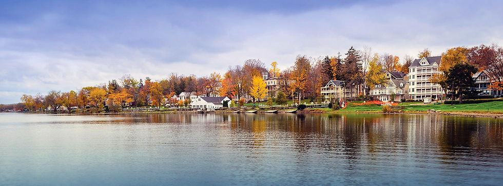 A panoramic landscape view across the placid waters of a large lake under a cloudy, overcast sky. On the far bank, a historic waterfront village featuring a collection of multi-story buildings, charming houses, and a large inn is nestled among mature trees displaying peak autumn foliage in vibrant golds and oranges. The calm lake surface reflects the buildings and the muted sky.