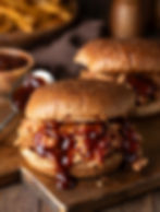 Two tender BBQ pulled pork sandwiches served on toasted buns with a generous drizzle of dark barbecue sauce, presented on a rustic wooden cutting board at Baseline Bar & Grille.