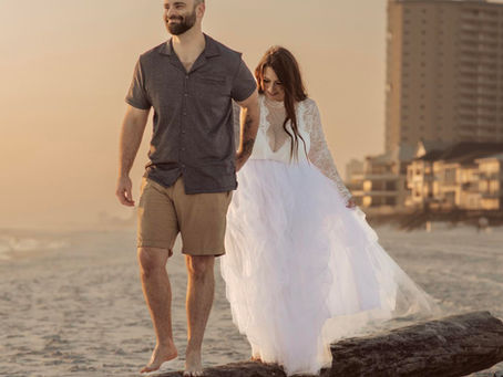 A couple walks barefoot on a log at a beach during sunset. The man wears a gray shirt and shorts, the woman a white wedding dress. Buildings in background. Elopement blog post.