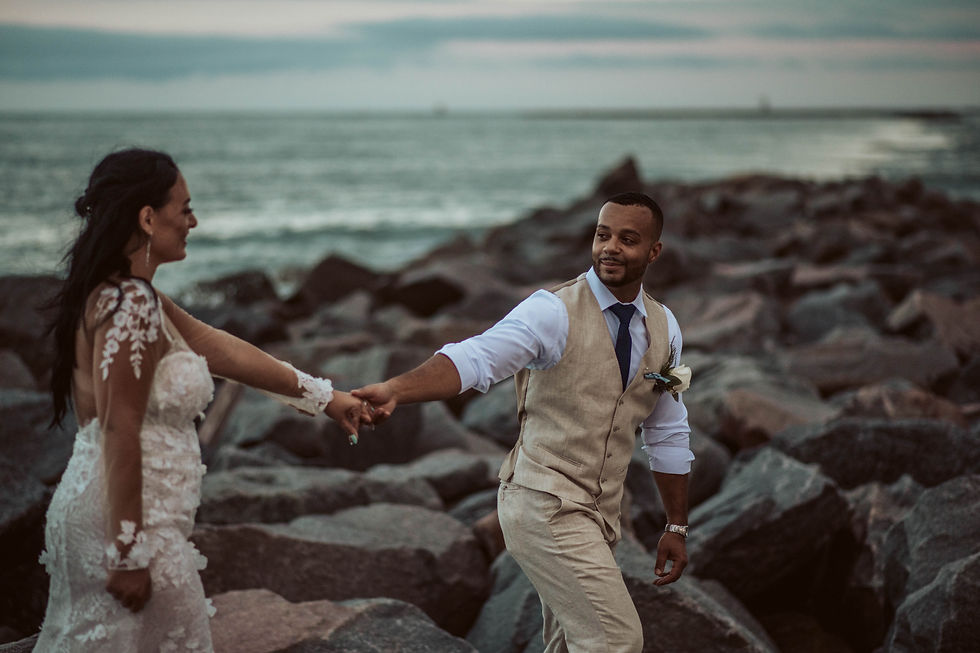 Couple in wedding attire walking on rocky shoreline at dusk, holding hands and smiling. Ocean waves and cloudy sky in background. Romantic mood.
