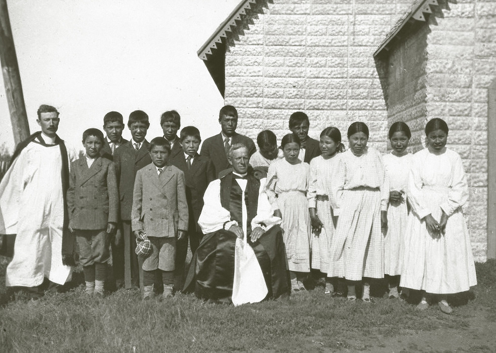 Archival black-and-white group photograph of Indigenous youth in suits and long dresses posing outdoors with two clergy members in robes.