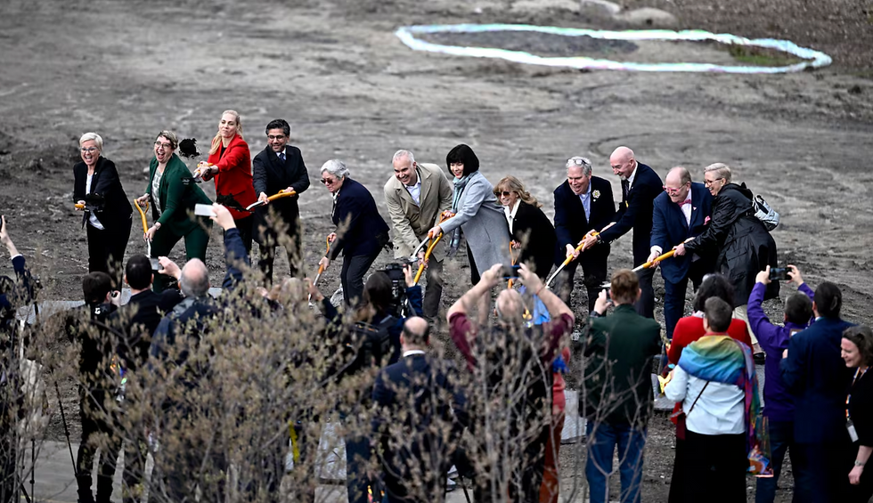 A group of people lined up outdoors using ceremonial shovels to break ground together during an official ceremony.
