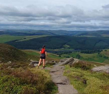 Rachel can be seen in the distance running in the peak district. lots of green hills can be seen in the background and a rocky path leads the way