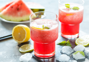 zwei Gläser mit rotem Erfrischungsgetränk, Zitrone und Eiswürfel. Dahinter ein Teller mit Melonenstücke