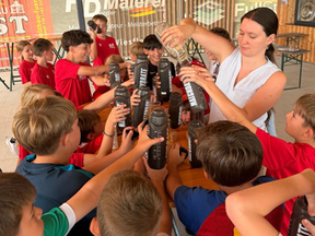 eine Frau teilt Getränke an eine Gruppe von Kindern aus, die ihre Wasserflasche hinhalten