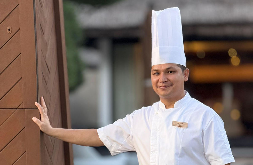 Smiling Chef Shyam Thapa in white uniform and tall hat leans on a wooden wall, with a blurred resort background.