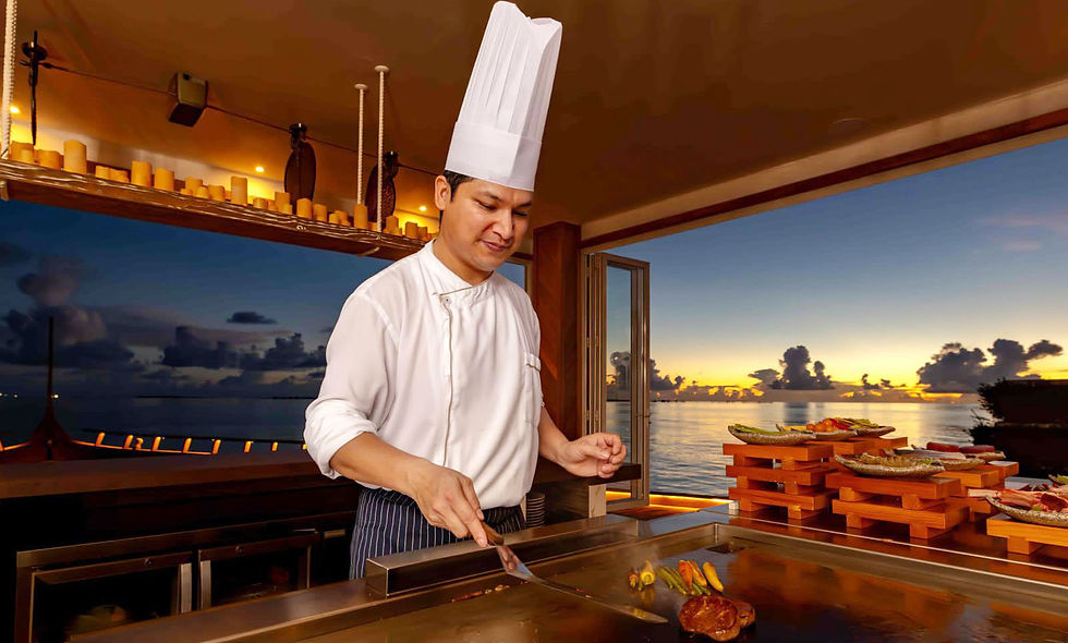 Chef Shyam Thapa in white hat cooks at a teppanyaki grill by the ocean at sunset, with plated food and a warm, calm mood.
